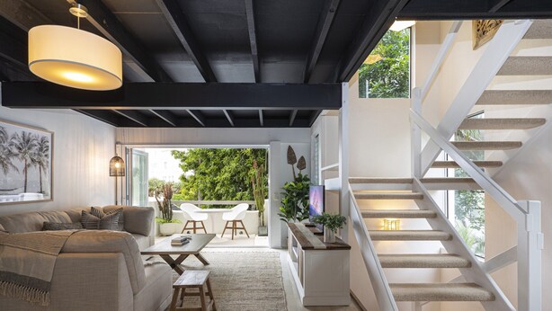 Ein modernes Wohnzimmer mit Sofa, Couchtisch und Sesseln, einer Treppe mit Geländer und Blick auf Grünfläche im Freien.
