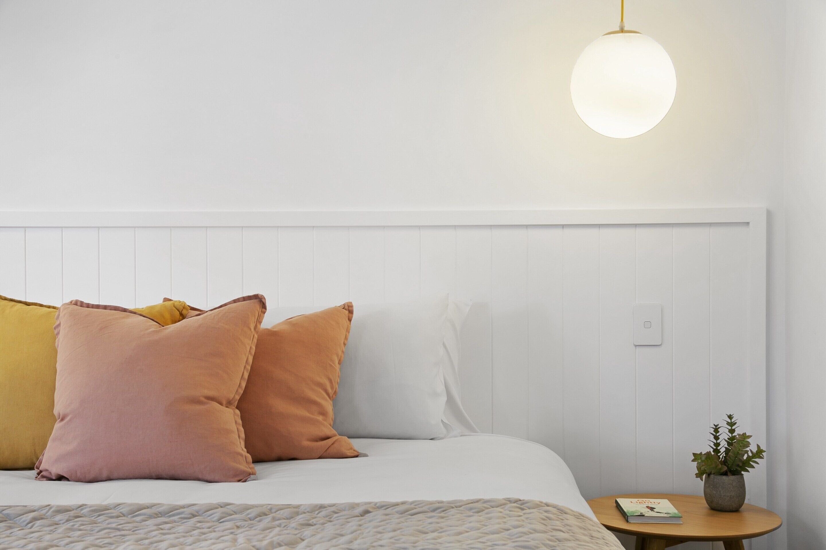 A neatly made bed with white and peach pillows, a round pendant light, and a small table with a plant and book.