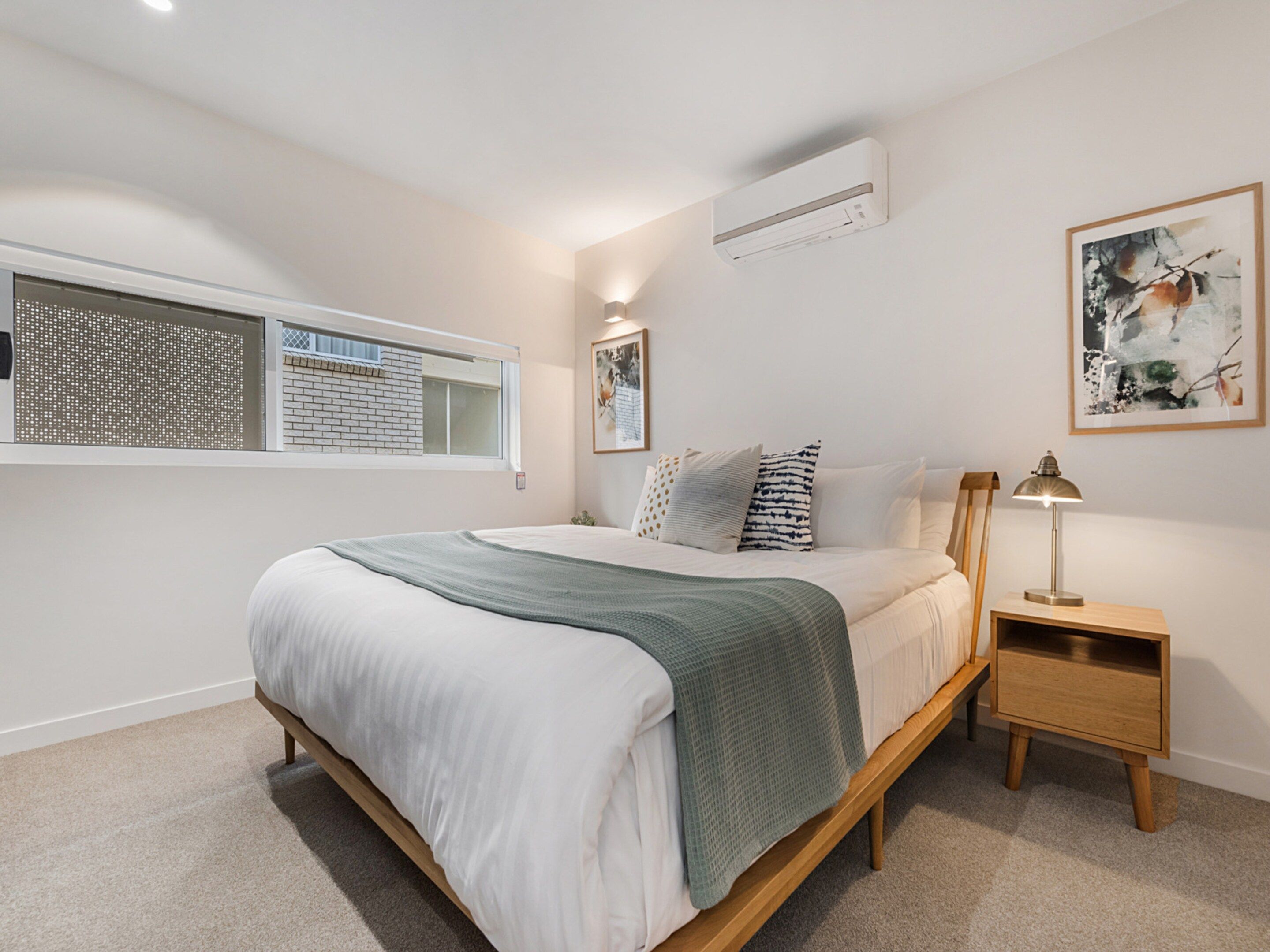 A bedroom with a bed, a nightstand, a lamp, a window, and an air conditioning unit.