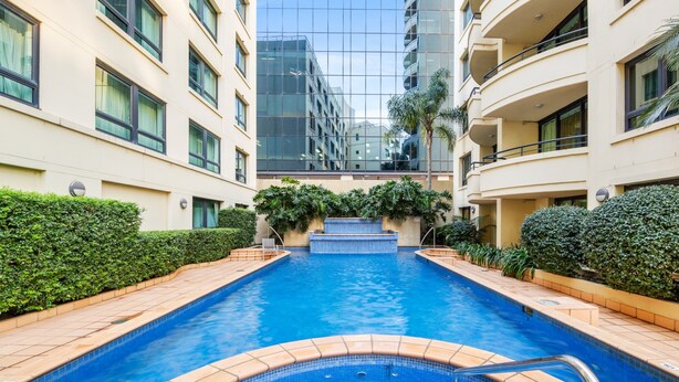 Una piscina con bordes de azulejos, rodeada de setos cuidados y un edificio con amplias ventanas y balcones.