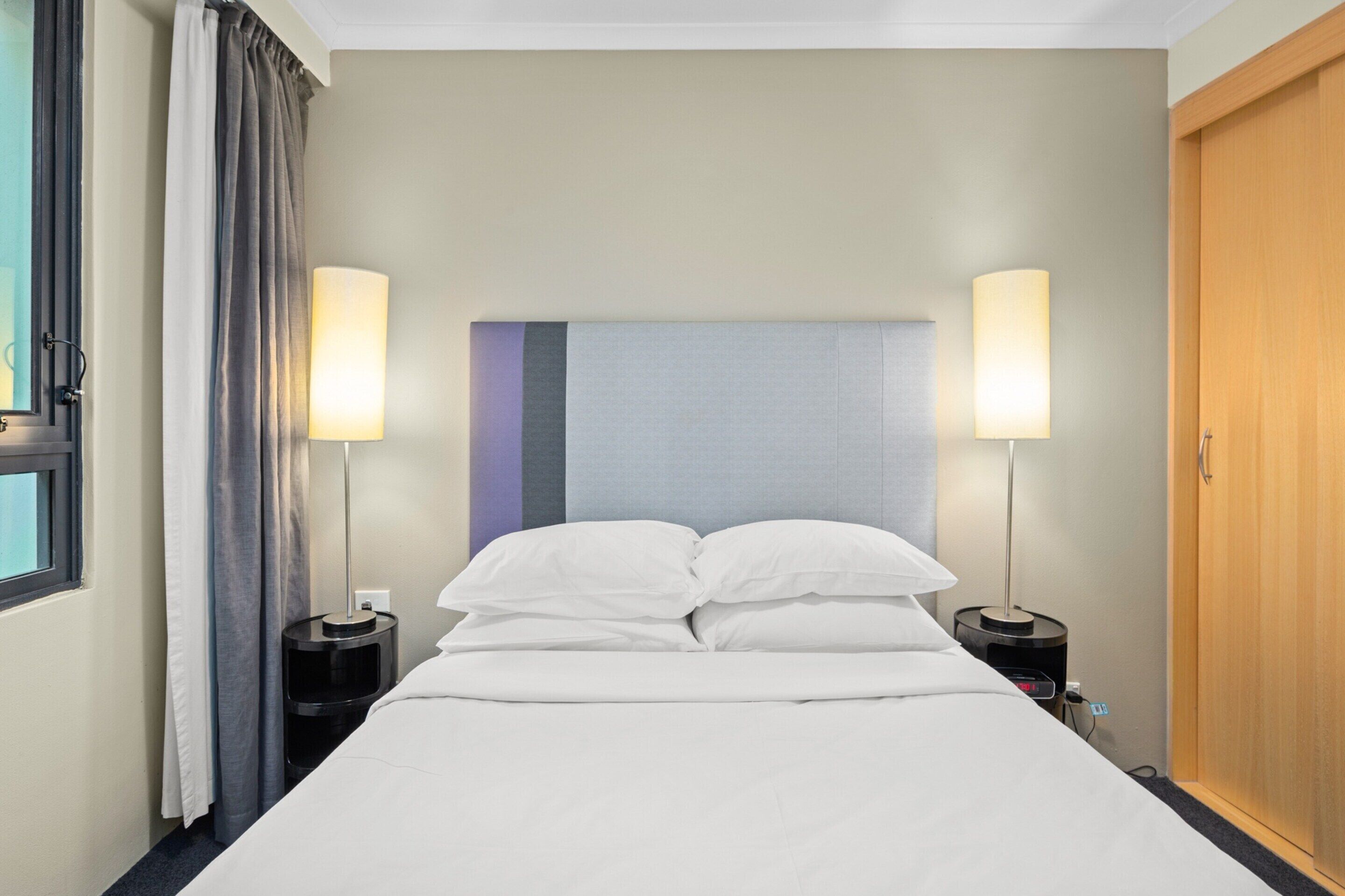Una habitación de hotel con una cama, dos lámparas de noche, una mesita de noche, una ventana con cortinas y una puerta de madera.