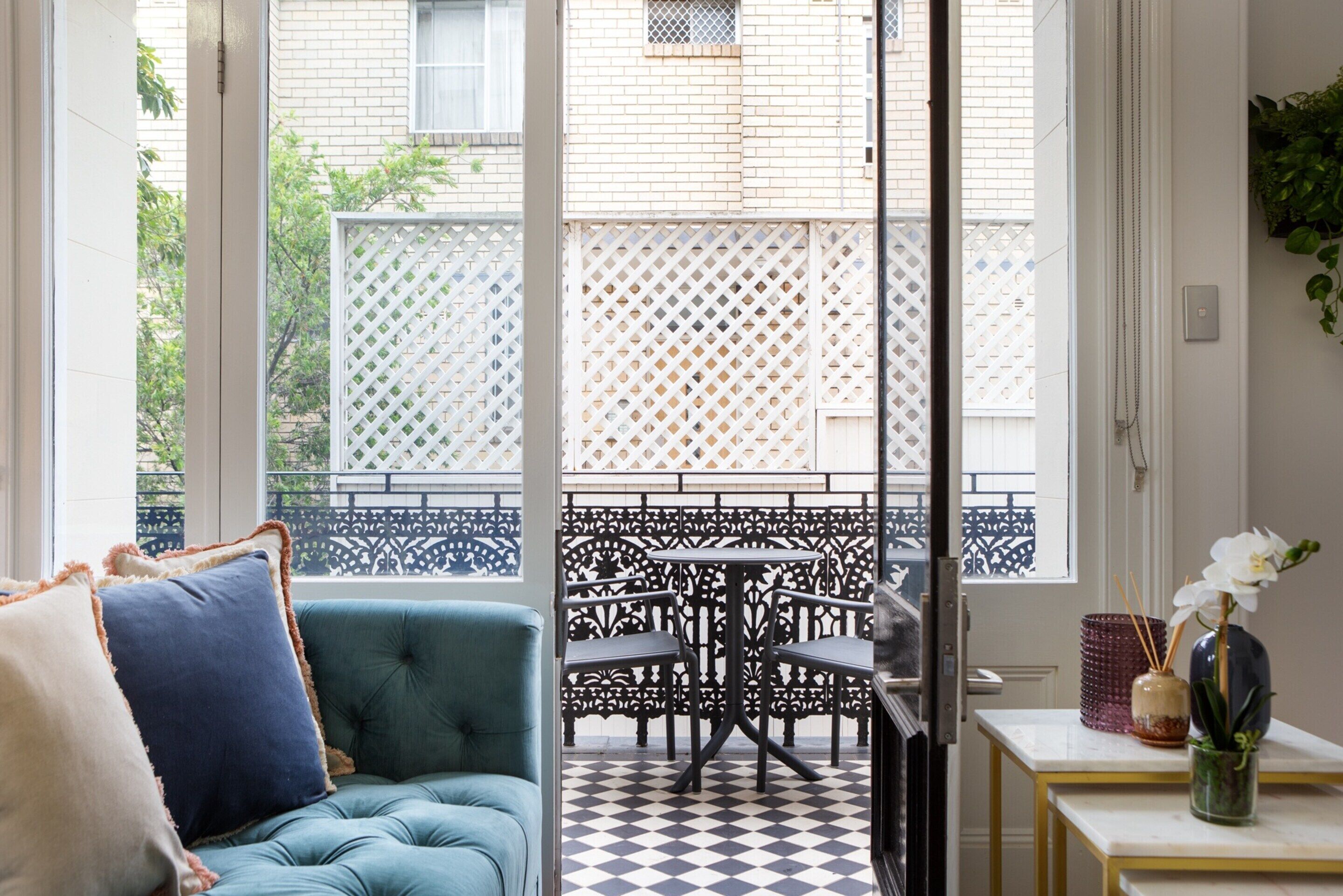 Une pièce avec un canapé de couleur turquoise, une petite table ornée d’un vase et d’une bougie, et une vue sur un balcon doté d’une balustrade en treillis.