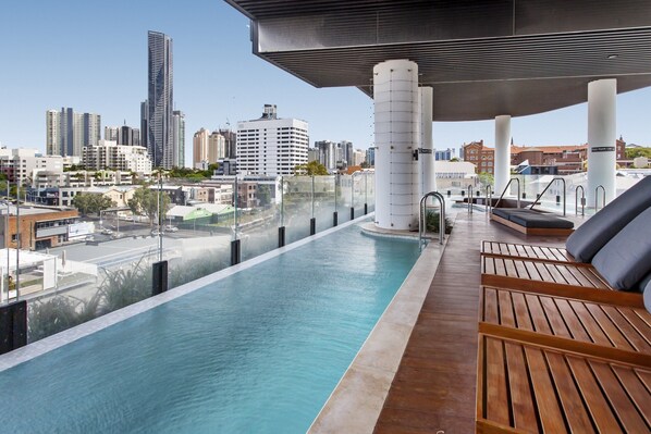 Piscina en la azotea con vistas a la ciudad, tumbonas de madera y una zona cubierta con columnas.