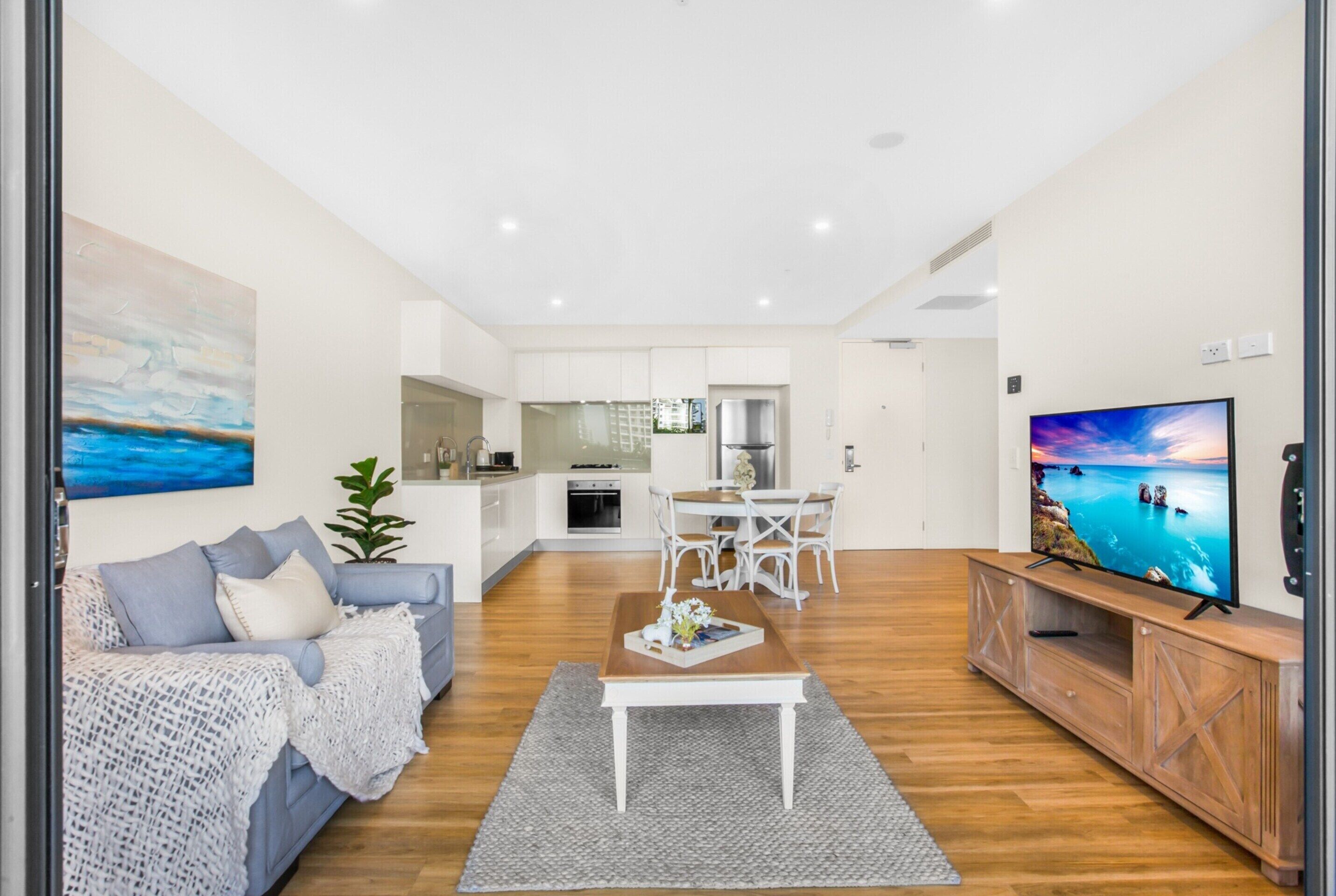 A modern living room with a sofa, coffee table, and TV. A kitchen is visible in the background.