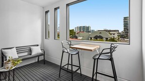 A balcony with a wooden table and two bar stools, a black bench with cushions, and a view of buildings through large windows. - Stellar Location With Parking, Pool & Gym Access (Brisbane)