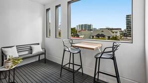 A balcony with a wooden table and two black bar stools, a black bench with white cushions, and a view of buildings through large windows. - Stellar Location With Parking, Pool & Gym Access (Brisbane)