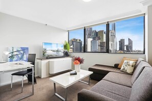 A modern living room with a white desk, a grey sofa, and a large window offering a city view. - Charming 1-bed Unit in an Unbeatable Location (Brisbane)