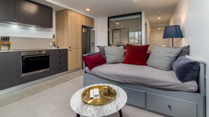 Una sala de estar moderna con un sofá gris, almohadas rojas y grises, una pequeña mesa redonda de centro con una bandeja, y una cocina con gabinetes oscuros y un horno empotrado.