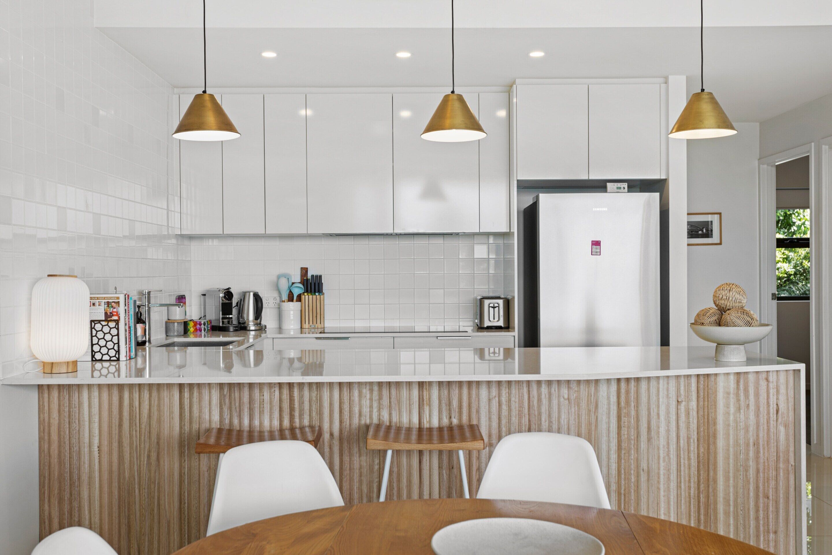 Uma cozinha moderna com armários brancos, uma ilha central com banquetas e iluminação pendente.