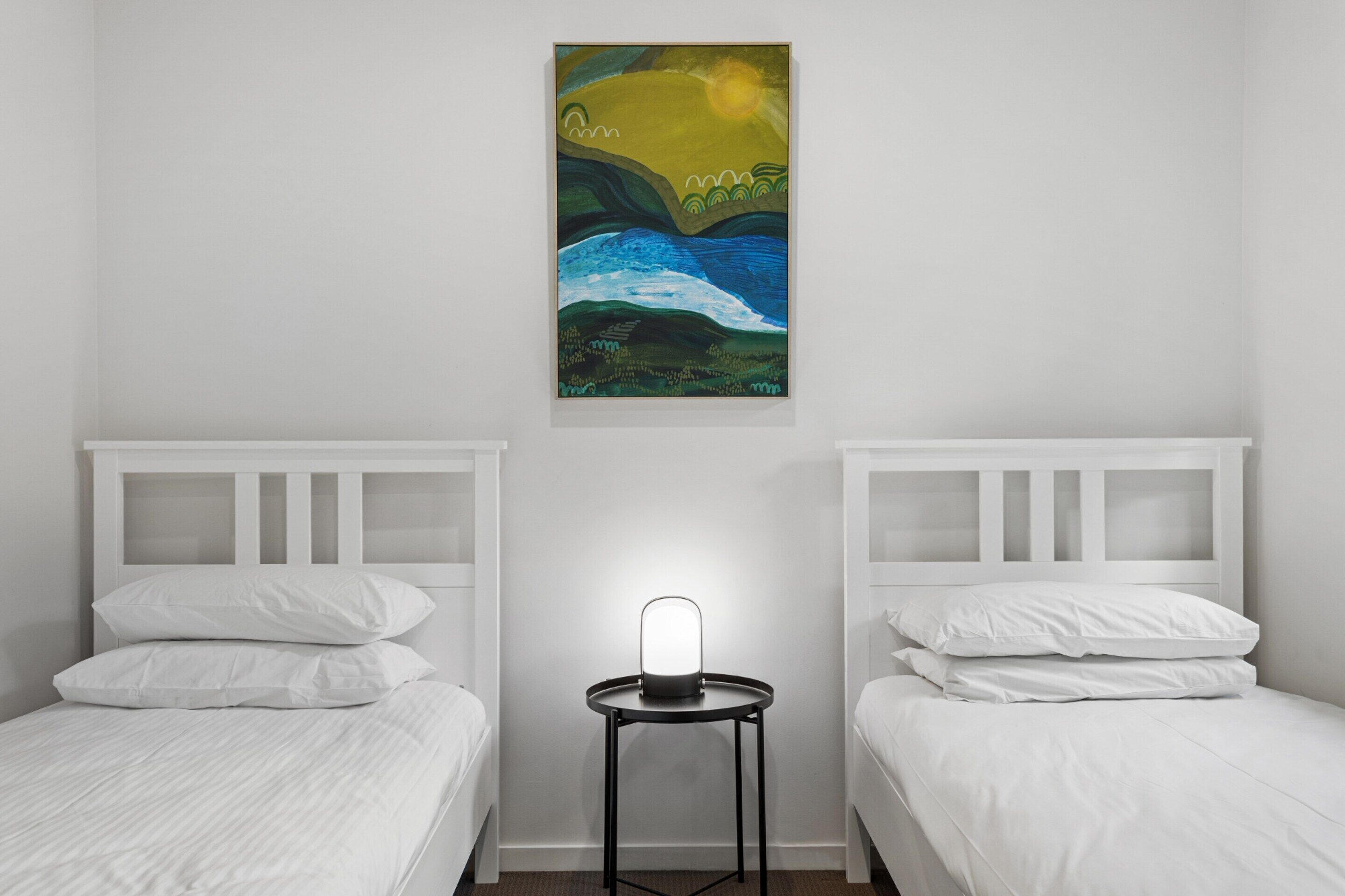 A hotel room with two single beds, a bedside table with a lamp, and a painting on the wall.