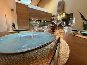 Una mesa de comedor dispuesta con platos, cubiertos y copas de vino, con una cocina moderna al fondo.