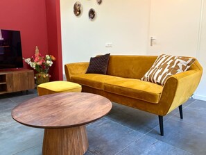 A living room with a mustard yellow sofa, a round wooden coffee table, and a yellow ottoman. - Zilverspar | Sfeervolle bosvilla met barrelsauna | Putten, Veluwe (Putten)