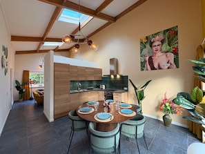 A modern kitchen with wooden cabinets, a dining area with a round table set for six, and a large window providing natural light. - Zilverspar | Sfeervolle bosvilla met barrelsauna | Putten, Veluwe (Putten)