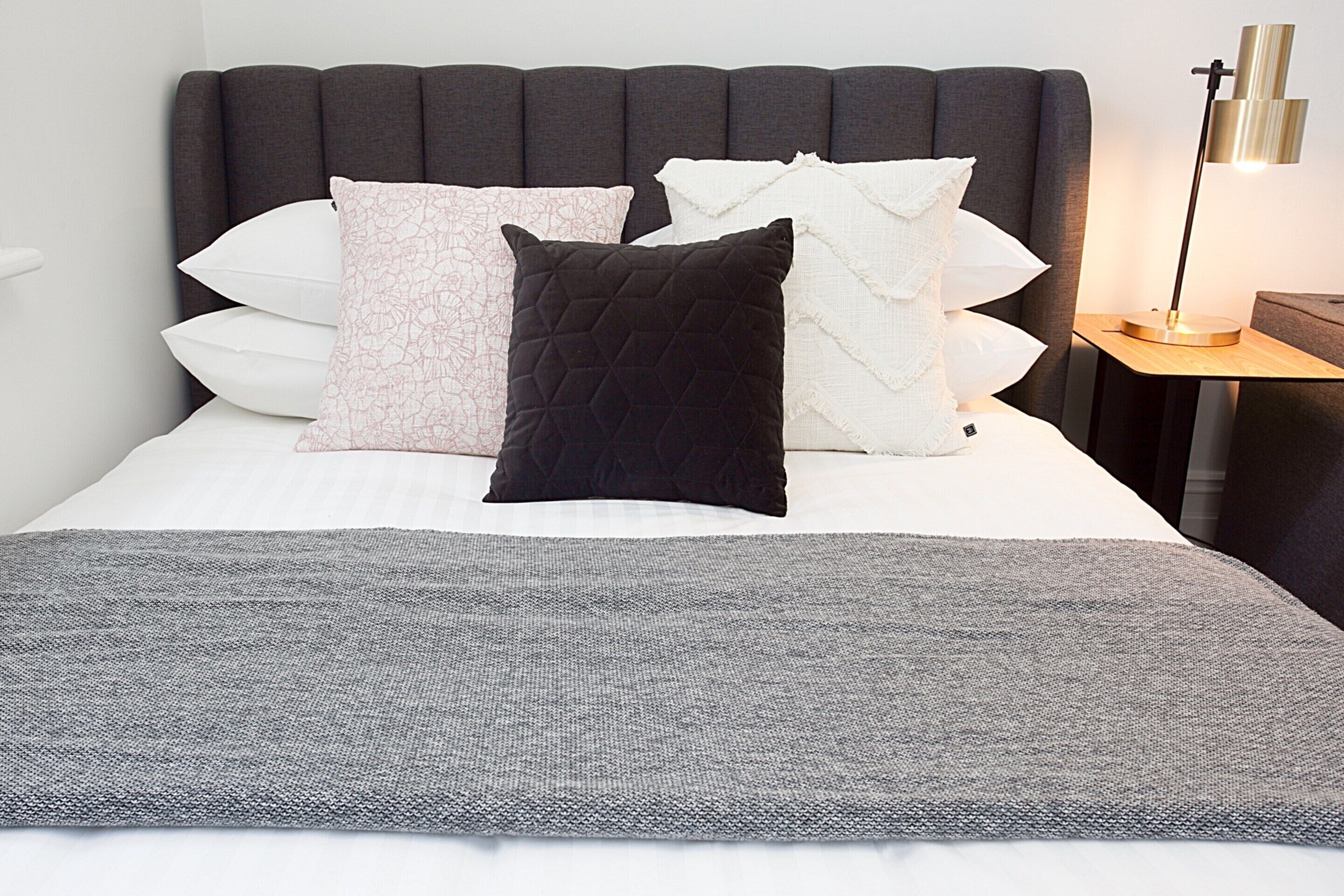 A neatly made bed with a grey blanket, a variety of pillows, and a bedside lamp on a wooden nightstand.