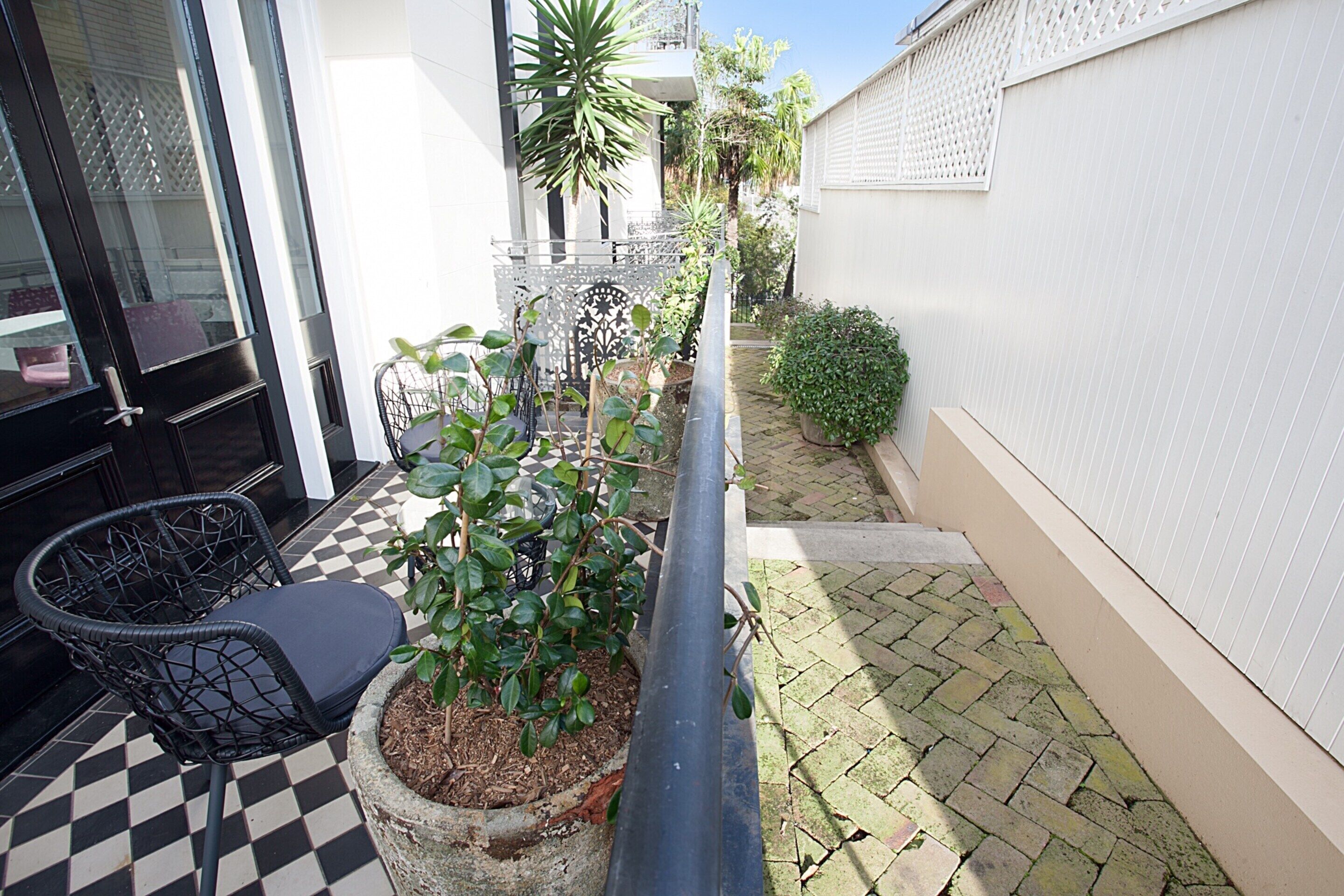 Un petit espace extérieur doté d’un sol à carreaux, d’une plante en pot, d’une chaise et d’un étroit chemin menant à une porte.