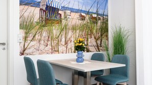 Een zithoek met een tafel en stoelen, een schilderij met een strandthema en een vaas met gele bloemen.