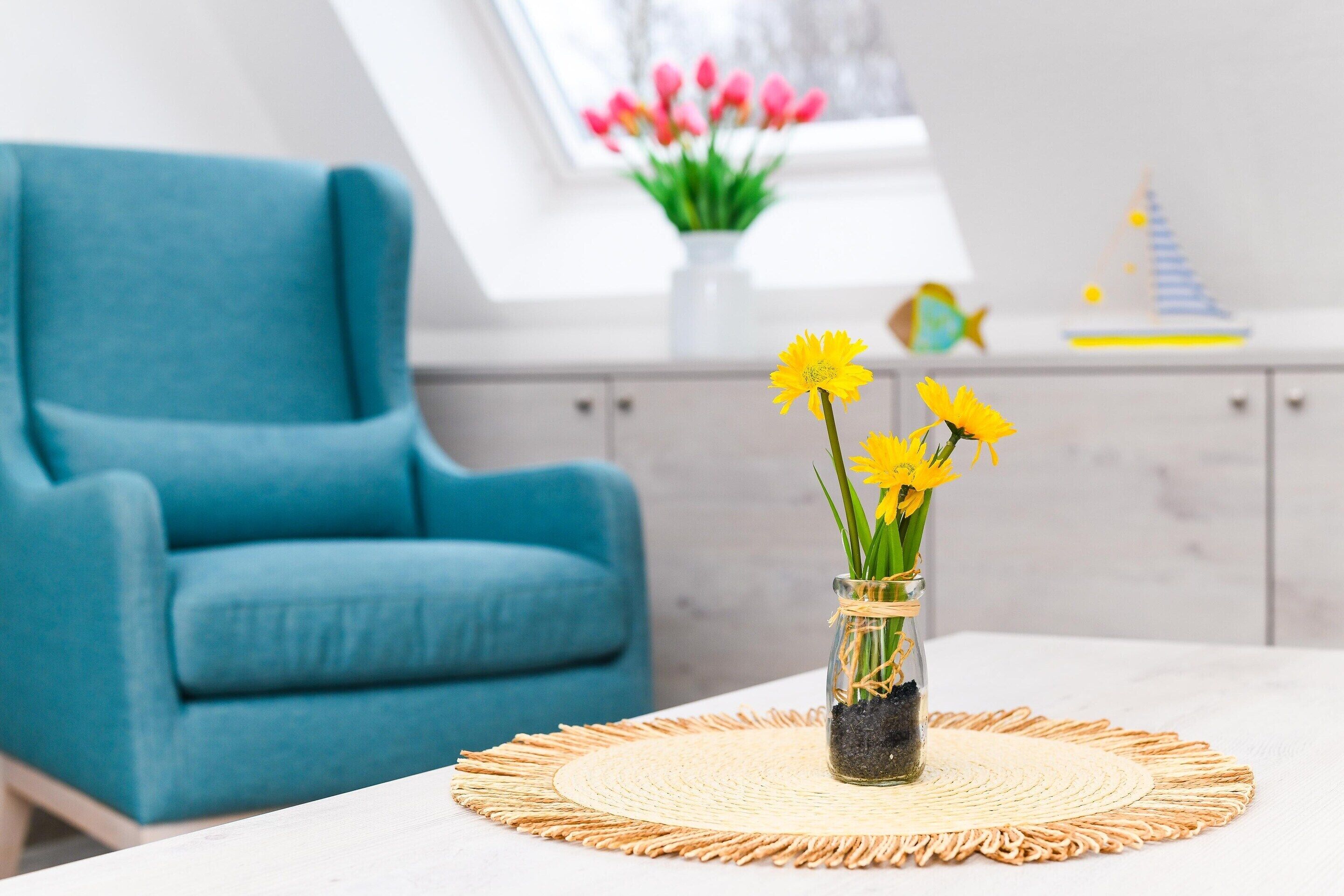 Et værelse med en blå lænestol, et bord med en vævet dug og en vase med gule blomster.