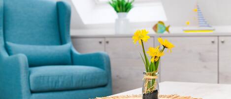 Et værelse med en blå lænestol, et bord med en vævet dug og en vase med gule blomster.