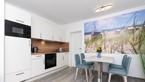 Een moderne keuken met witte kasten, een zwarte oven en een houten aanrecht. Er is een zithoek met een witte tafel en twee blauwgroene stoelen. Op de muur hangt een schilderij van een strandgezicht.