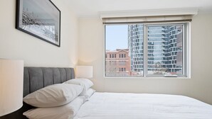 A neatly made bed with white linens and pillows, a gray upholstered headboard, a bedside lamp, and a framed picture on the wall. - Gorgeous 3-bed Apartment Near Flagstaff Gardens (Melbourne)