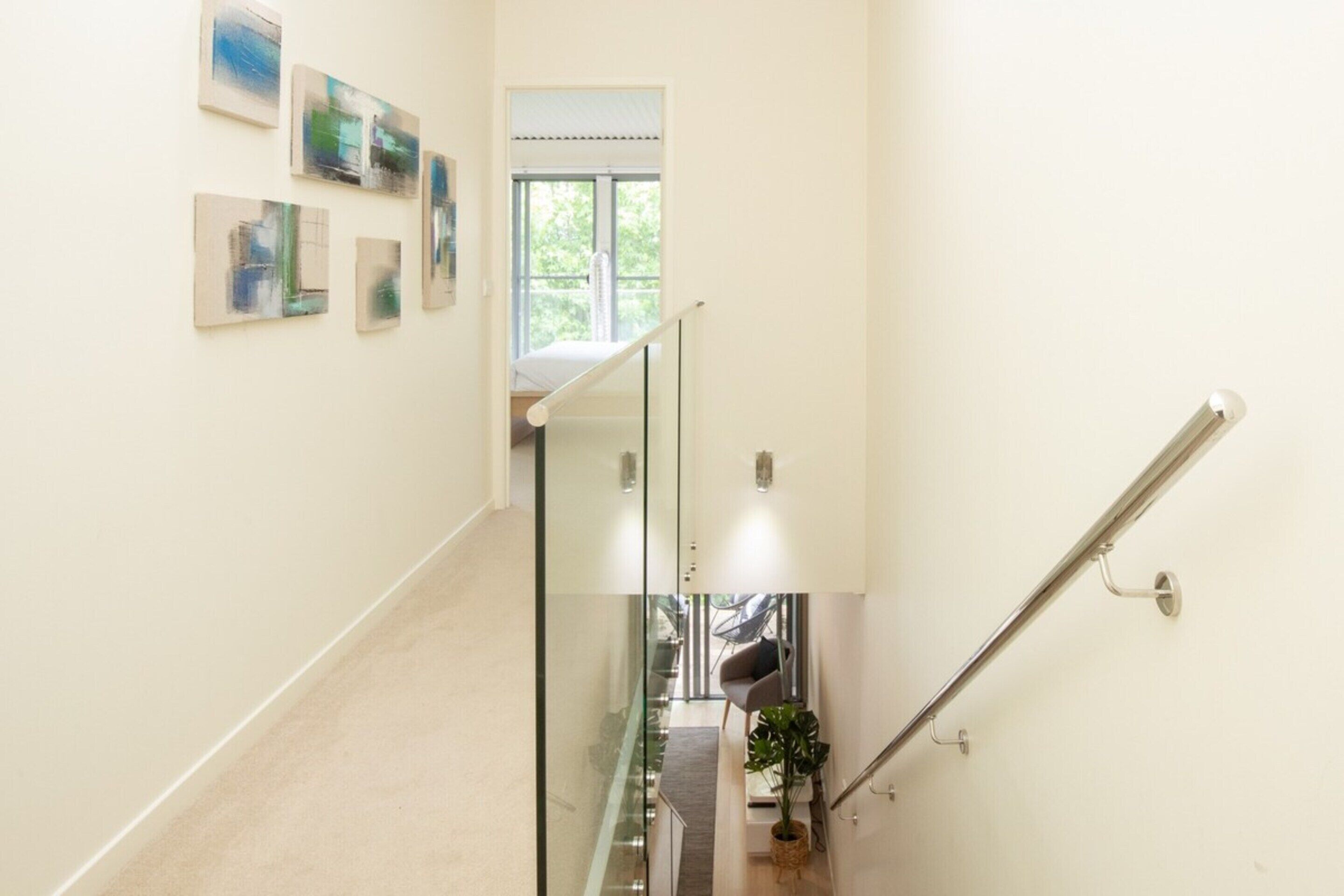 Una moderna scala con ringhiere in vetro, vista su una stanza con un letto e una pianta, e arte astratta al muro.