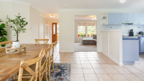 A spacious kitchen with white cabinets, a dining area with a wooden table and chairs, and a view of a living room through an open doorway. - Stunning Katoomba Home With Lush Garden&playground (Katoomba)