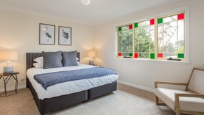 A bedroom with a bed, two bedside lamps, a nightstand, a window with colored panes, and two framed pictures on the wall. - Stunning Katoomba Home With Lush Garden&playground (Katoomba)