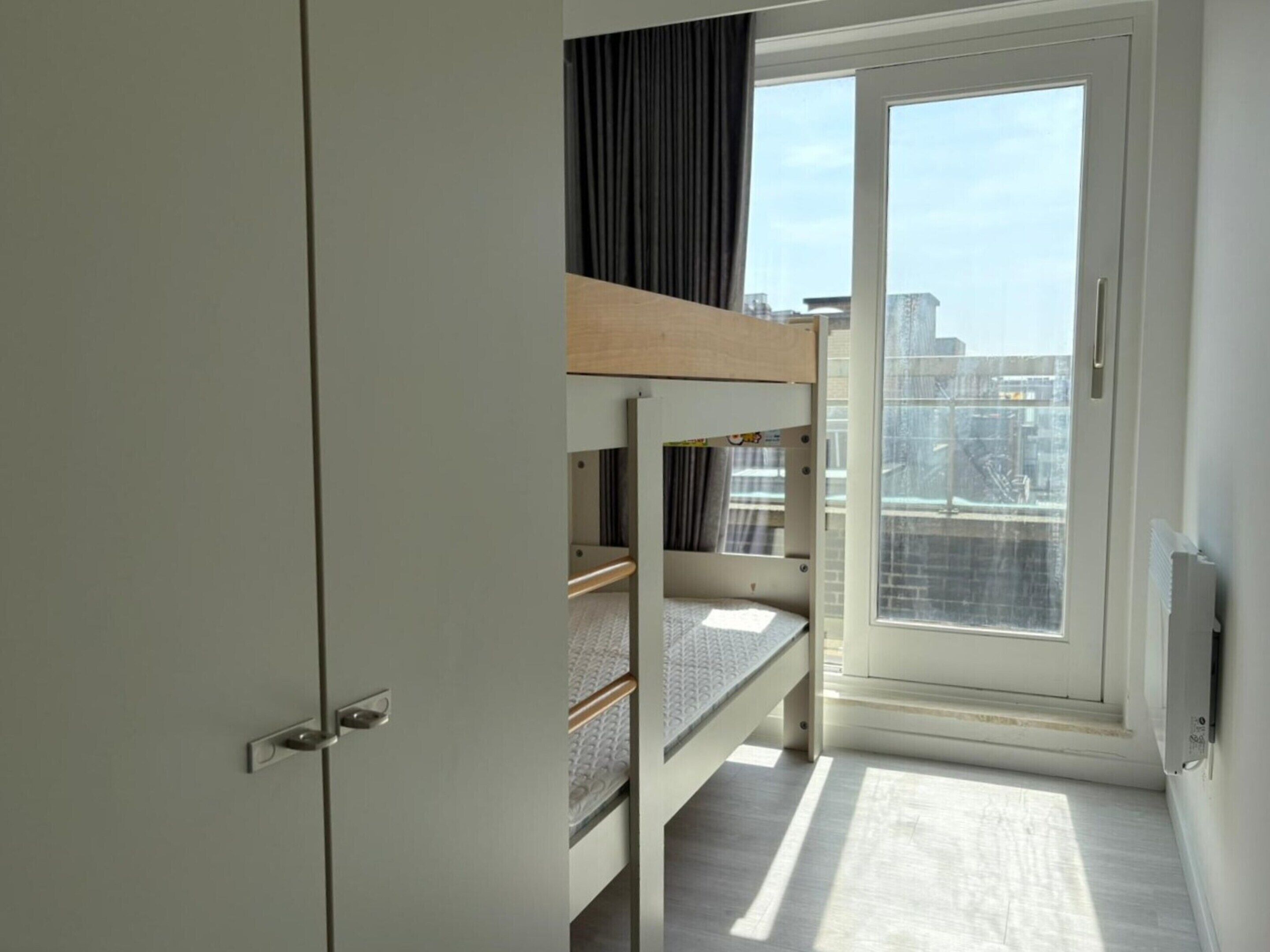 A small, modern room with a bunk bed, a sliding door to a balcony, and a view of the cityscape.