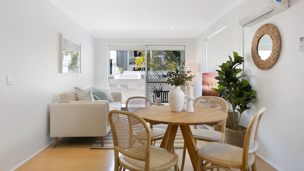 A modern living room with a white sofa, wicker chairs, a wooden dining table, and a large window with a view of a balcony and outdoor plants. - Ground Floor 2-bed Mermaid Beach Pad With Parking (Gold Coast)