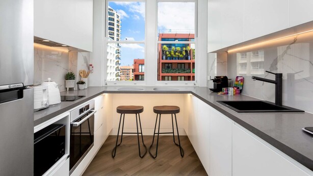 A modern kitchen with two bar stools, a built-in oven, and a window with a city view. - Stylish Studio Near Kings Cross Train Station (Sydney)