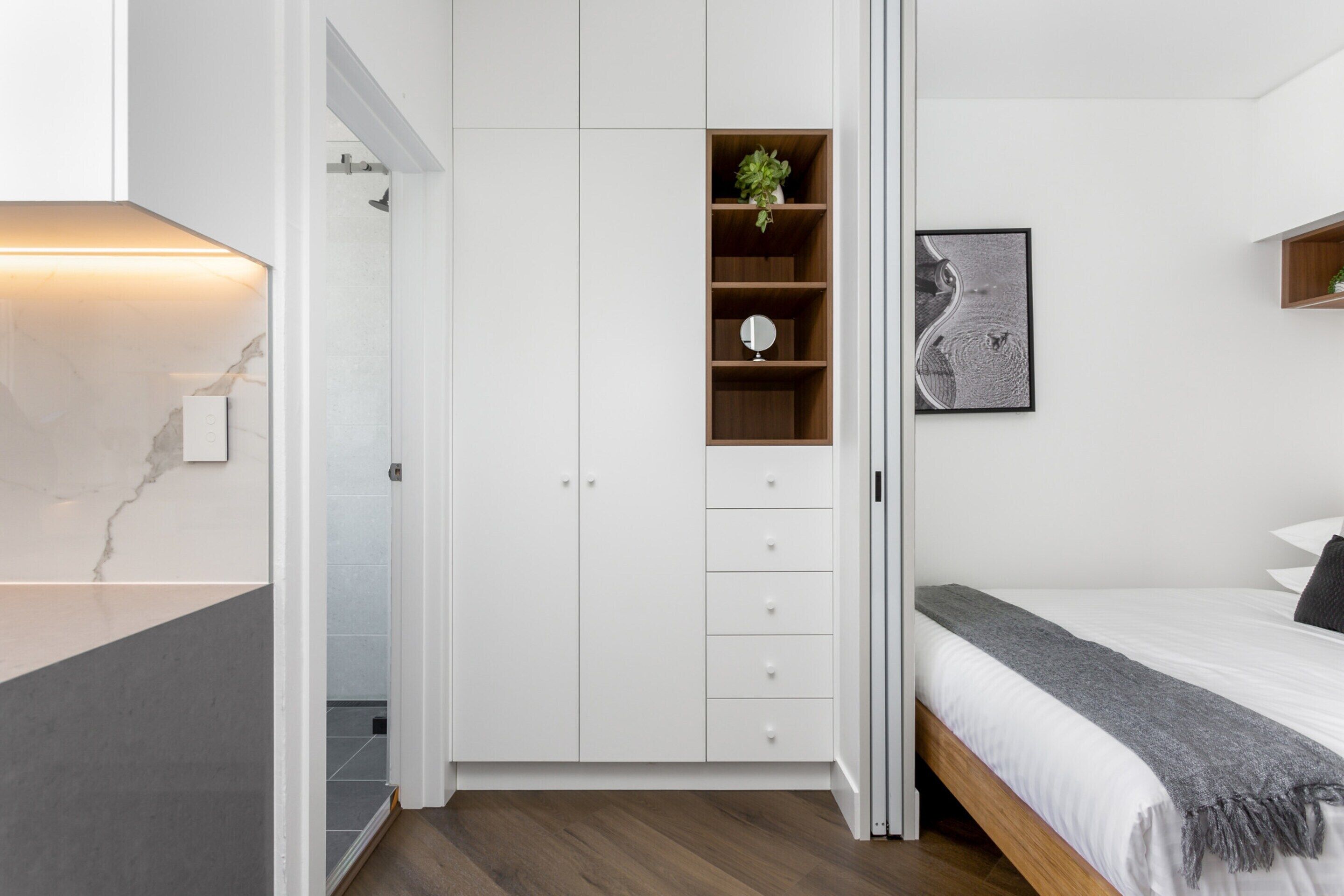 Ein modernes Schlafzimmer mit einem Bett, einem Schrank mit Schubladen, einem kleinen Regal mit einer Pflanze und einem gerahmten Bild an der Wand.