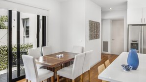 A modern dining area with a wooden table and white chairs, a blue vase, and a view of the outdoors through large windows. - 2-bed Apartment With Parking & Balcony Near Trains (Melbourne)
