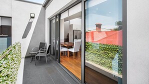 A modern patio with a dining area, a glass door leading to a kitchen, and a view of a garden. - 2-bed Apartment With Parking & Balcony Near Trains (Melbourne)