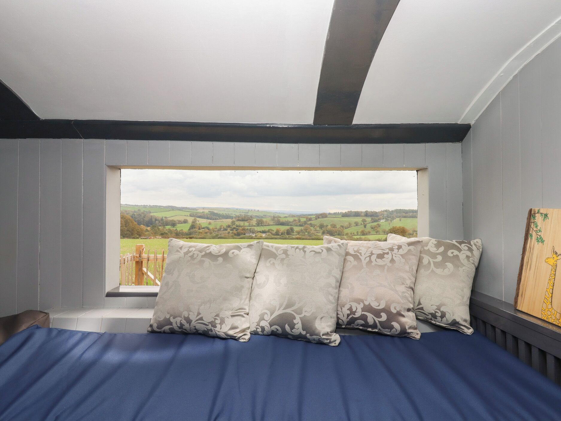 A bedroom with a large window, a bed with blue bedding, and three patterned pillows.