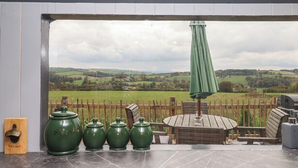 A patio area with a view of rolling hills, a wooden fence, and a green umbrella. - The Bus on the Hill (Tiverton)