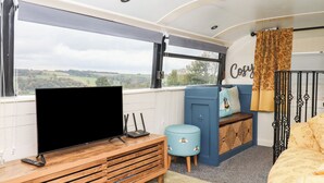 A cozy interior with a TV on a wooden stand, a blue cabinet, a small stool, and a bed with a floral bedspread. - The Bus on the Hill (Tiverton)
