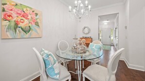 A dining area with a glass table, white chairs, a chandelier, and a painting of flowers on the wall. - Pool | Pet Friendly | Clean & Bright | Patio | Grill | Great Kitchen | Beach Chairs (St. Simons Island)