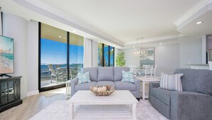 Uma sala de estar espaçosa com um sofá cinzento, uma mesa de centro branca e vista para a cidade através de grandes janelas.