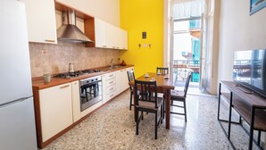 Cucina con mobili bianchi, un piano di lavoro in legno, un fornello a gas e una cappa aspirante in acciaio inossidabile. C'è un tavolo da pranzo con sedie e una TV su un mobiletto in legno.