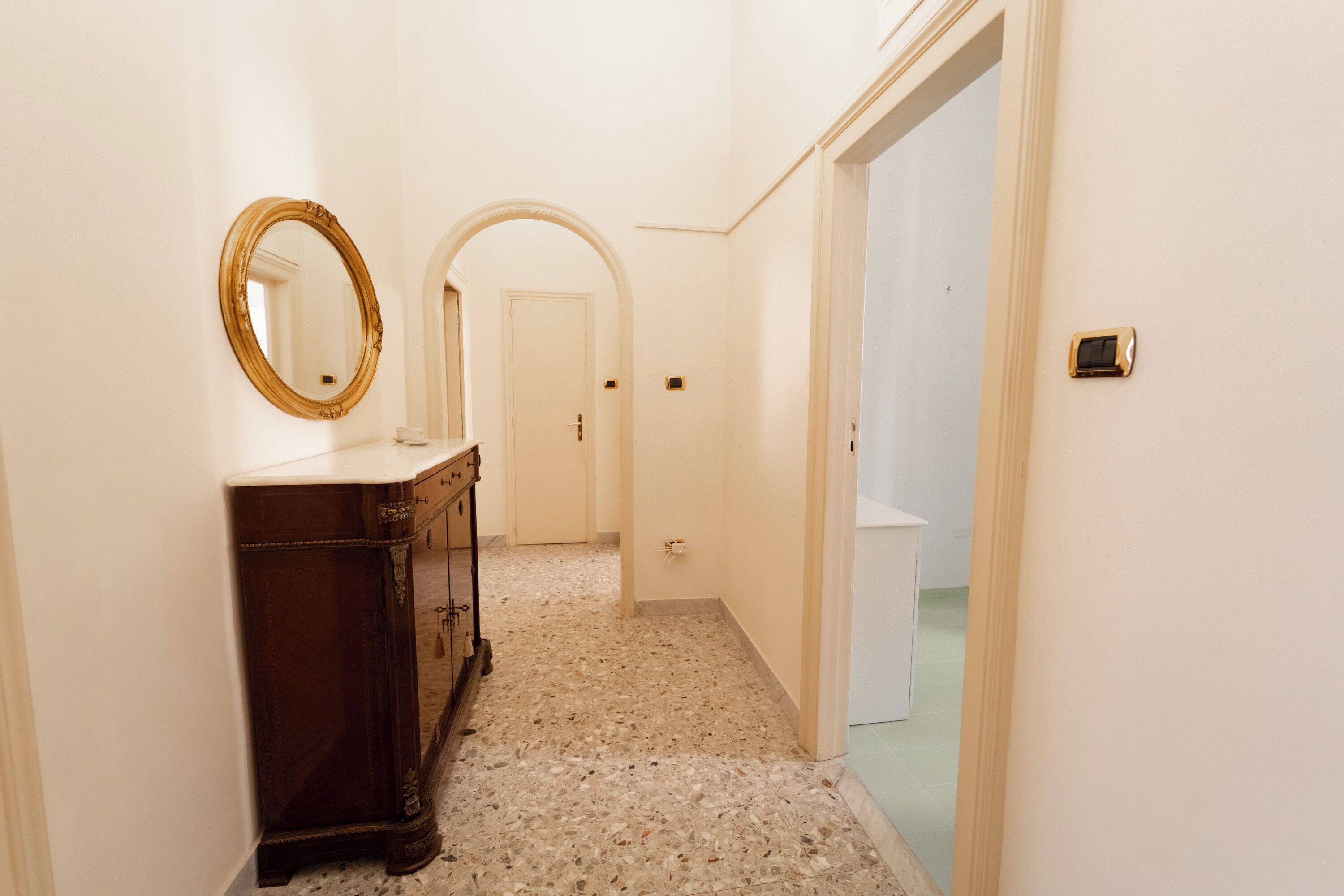 A hallway with a marble floor, a wooden dresser with a mirror, and a door at the end.