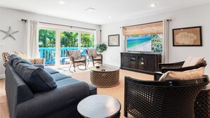 Un salon comprenant un canapé, des fauteuils, une table basse et un téléviseur. Il y a des fenêtres avec des rideaux et une vue sur l’extérieur.