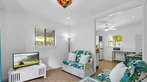 A living room with a television on a white console, a white wicker sofa with tropical cushions, a glass coffee table, and a ceiling fan. - The Little Blue Shack - Marcoola Granny Flat (Sunshine Coast)