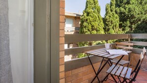 Un balcone con tavolo e sedie in legno, una tazza bianca e una finestra che si affaccia su un edificio in mattoni e alberi alti.