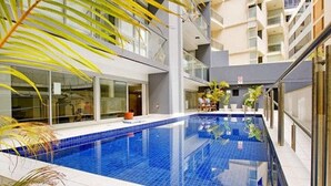A modern indoor swimming pool with clear blue water, surrounded by glass walls and balconies, and potted plants. - Stylish Studio With Pool & Gym in Heart of Sydney (Sydney)
