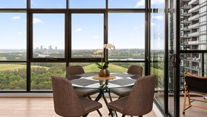 A modern dining area with a glass table and chairs, a vase with flowers, and a view of a cityscape through large windows. - Chic 1-bed With Pool by River, Ferry & Amenities (Sydney)