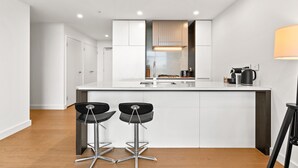A modern kitchen with white cabinetry, a central island with two bar stools, and a coffee machine. - Chic 1-bed With Pool by River, Ferry & Amenities (Sydney)