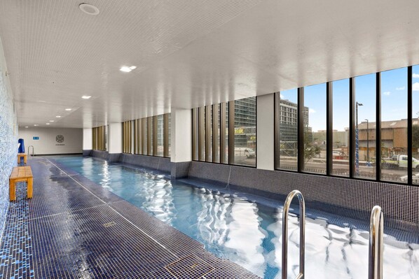 An indoor swimming pool with clear glass walls, a tiled floor, and a wooden bench. - Chic 1-bed With Pool by River, Ferry & Amenities (Sydney)