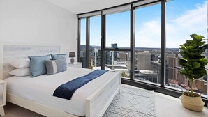 A modern bedroom with a large bed, a nightstand, a bedside table, a potted plant, and a city view through large windows. - Breathtaking City Views 1-bed With Pool, BBQ & Gym (Brisbane)