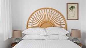 Un lit avec une tête de lit en rotin, du linge de lit blanc, deux lampes de chevet et une illustration botanique encadrée accrochée au mur.