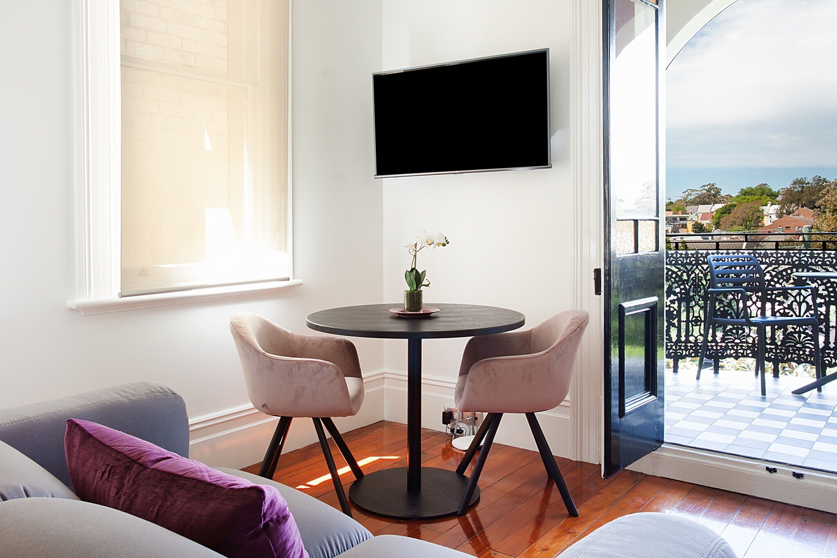 A modern room with a round table, two chairs, a TV mounted on the wall, and a balcony with outdoor furniture.